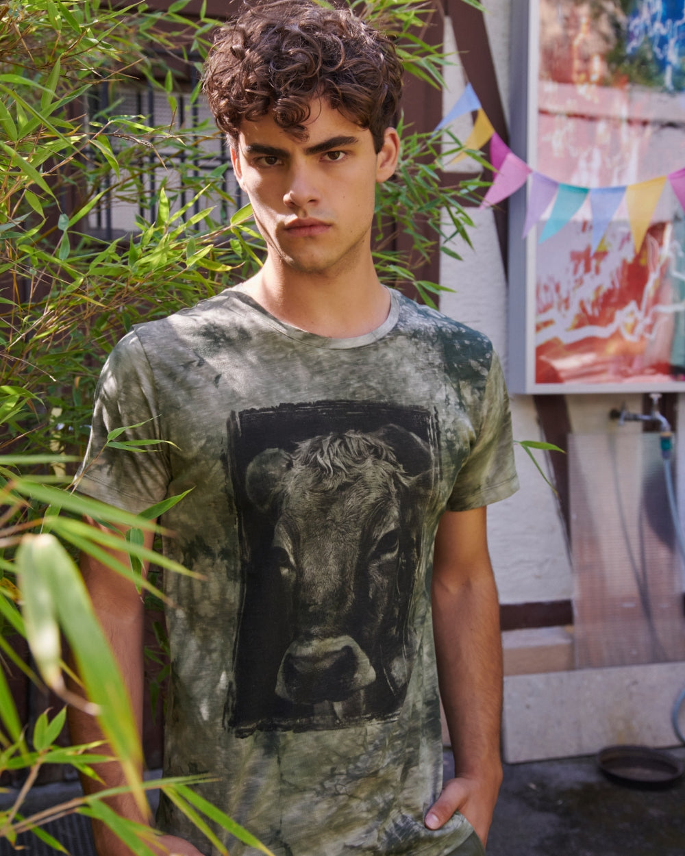 Model steht neben Schilfgrün und trägt ein grünes Batik Shirt mit dem Aufdruck eines Kuh Kopfes