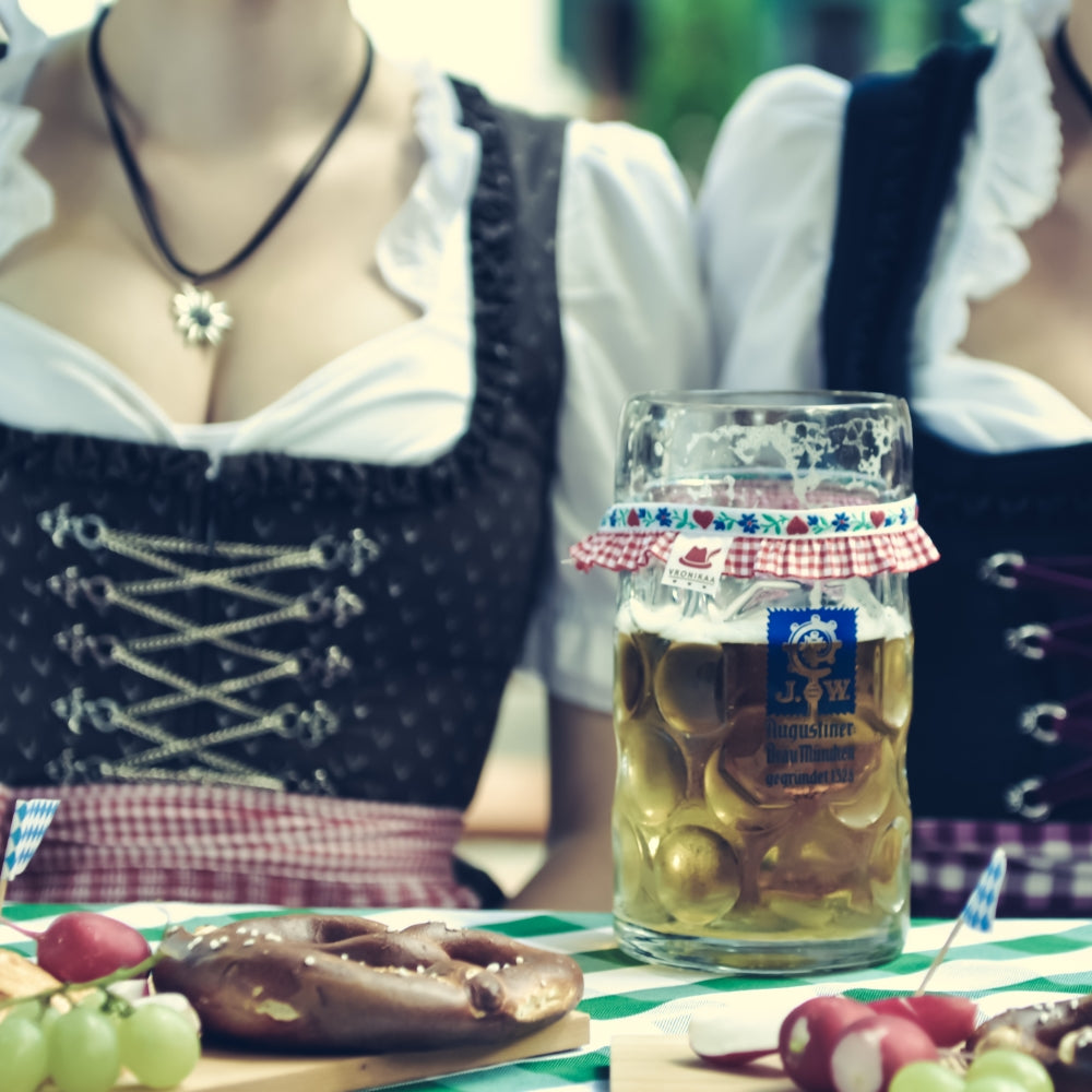 Maßkrug steht auf dem gedeckten Brotzeittisch und im Hintergrund sind zwei Dirndls