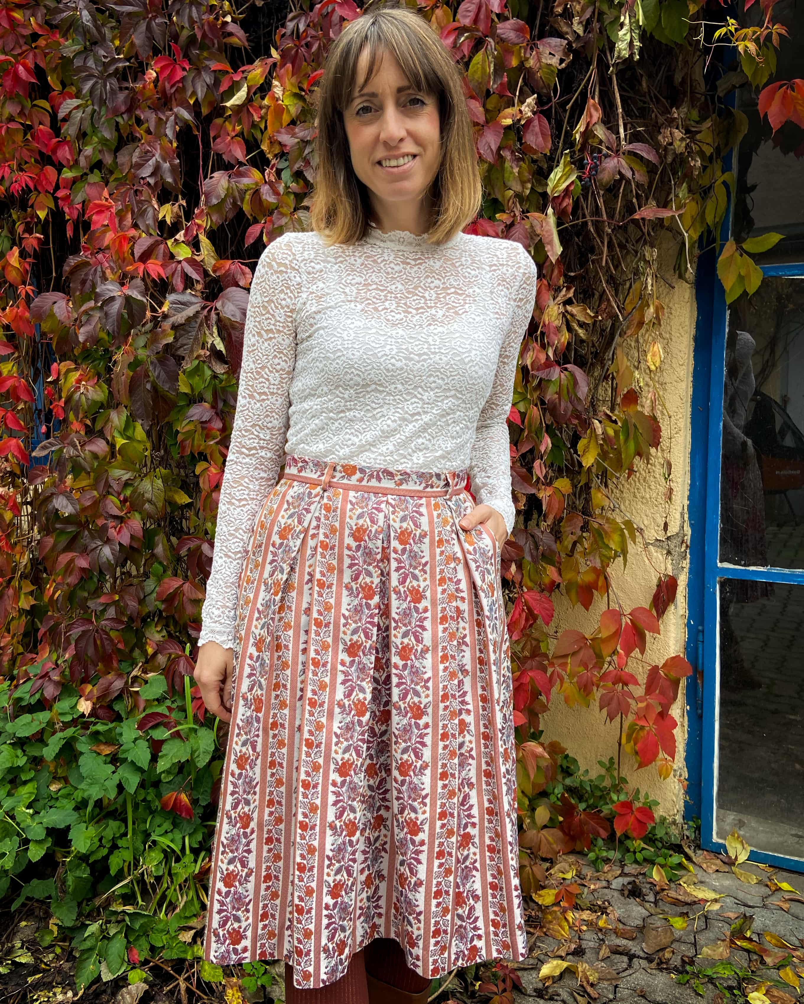 Model steht vor einer schönen Wand mit buntem Laub und trägt ein Spitzenshirt mit elegantem Trachtenrock