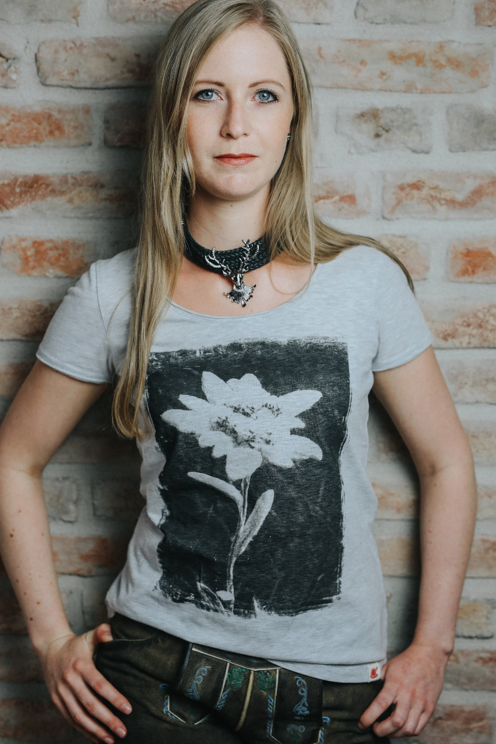 Weibliches Model mit blonden Haaren trägt Lederhose und das passende Trachtenshirt mit Edelweiß Foto-Motiv