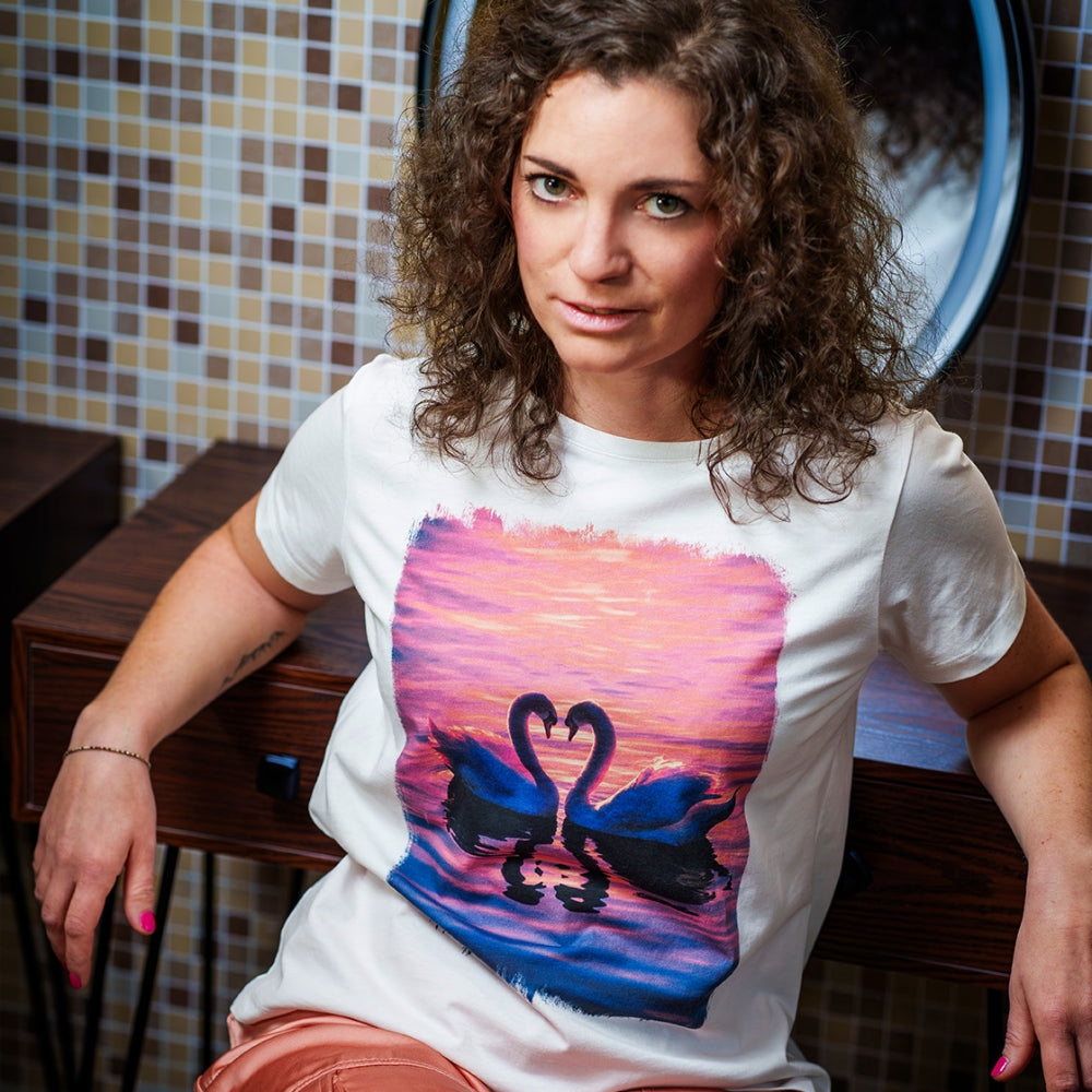 Model schaut in die Kamera und trägt ein T-Shirt mit einem Foto-Motiv von zwei Schwänen die mit ihrem Hals ein Herz bilden. 