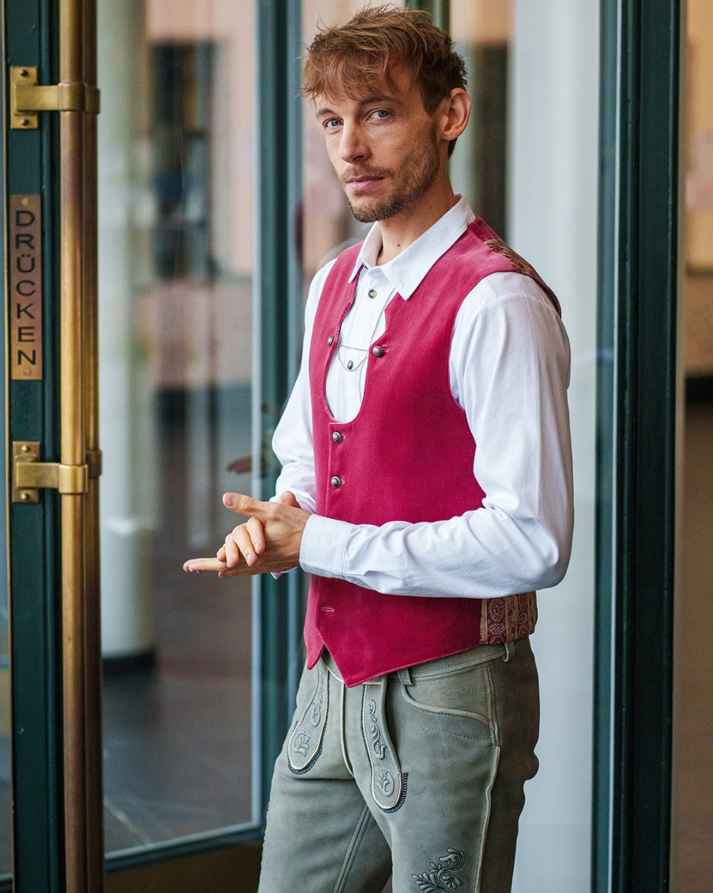 Ein Mann in Lederhose steht vor der Tür und schaut in die Kamera, er trägt ein Jersey Hemden Shirt in Weiß von vronikaa dazu eine rosa Weste