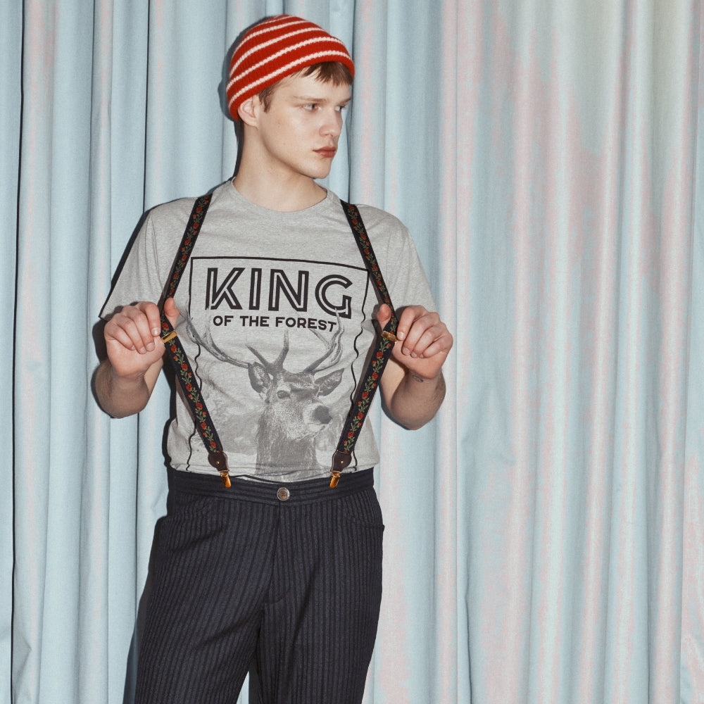 Model steht vor einem Vorhang mit roter Mütze, Hosenträgern und dem grauen Shirt mit großem Aufdruck eines Hirsches und dem Schriftzug King of the Forest