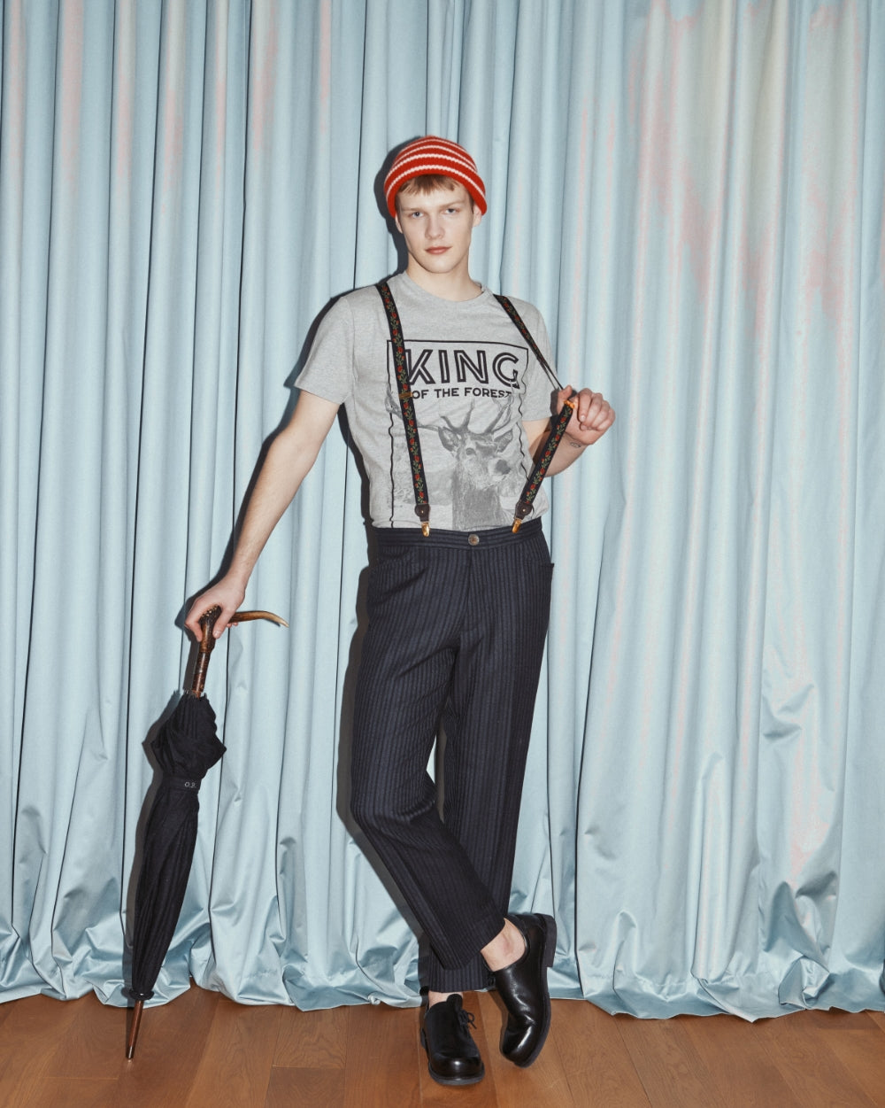 Model steht vor einem Vorhang und trägt Hose mit Hosenträgern und das graue Shirt mit großem Aufdruck eines Hirsches und dem Schriftzug King of the Forest