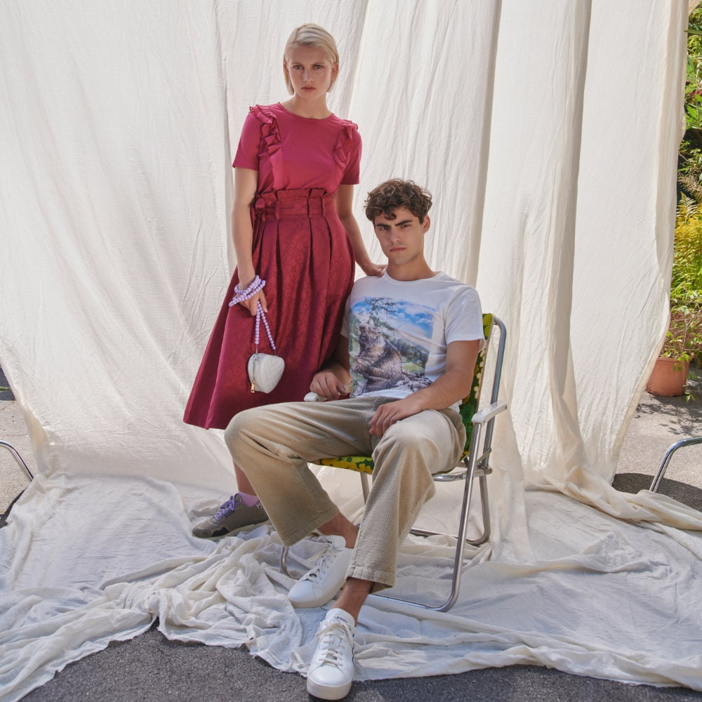 Zwei Models posieren vor einem hellen Vorhang, er sitzt auf einem Retro-Klappstuh und sie steht neben ihm in einem roten Outfit im Alpine Style